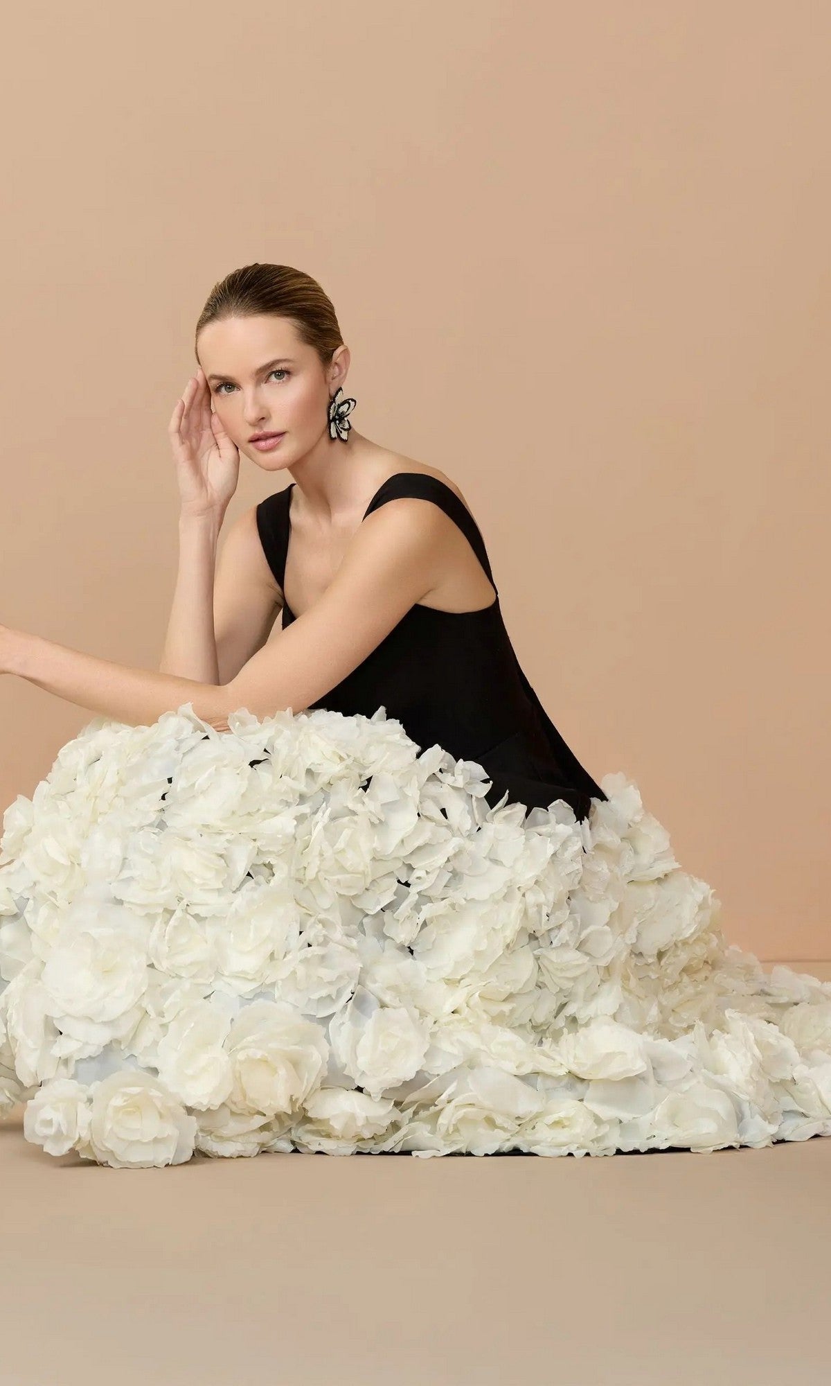 A woman poses seated against a beige background, wearing the Midi Wedding Guest Dress: Mac Duggal 21080—a black sleeveless top with a voluminous skirt adorned with dramatic 3D organza flowers.